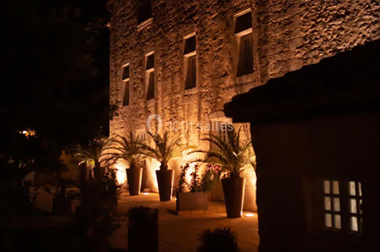 Façade en pierre éclairée de nuit, bordée de grands pots avec des palmiers et des fenêtres illuminées.