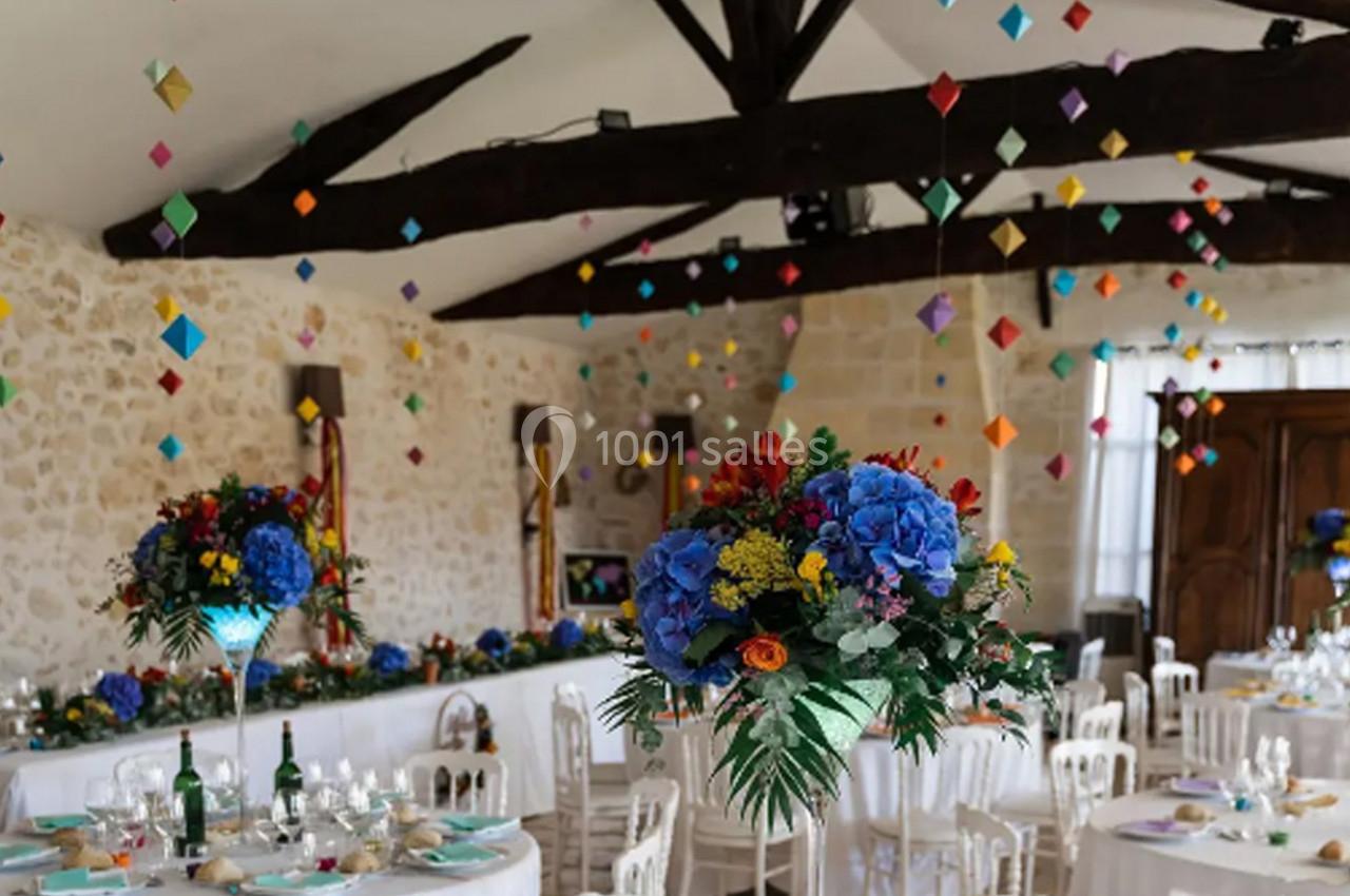 Salle de réception décorée avec des guirlandes colorées suspendues et des centres de table fleuris sur des tables dressées.