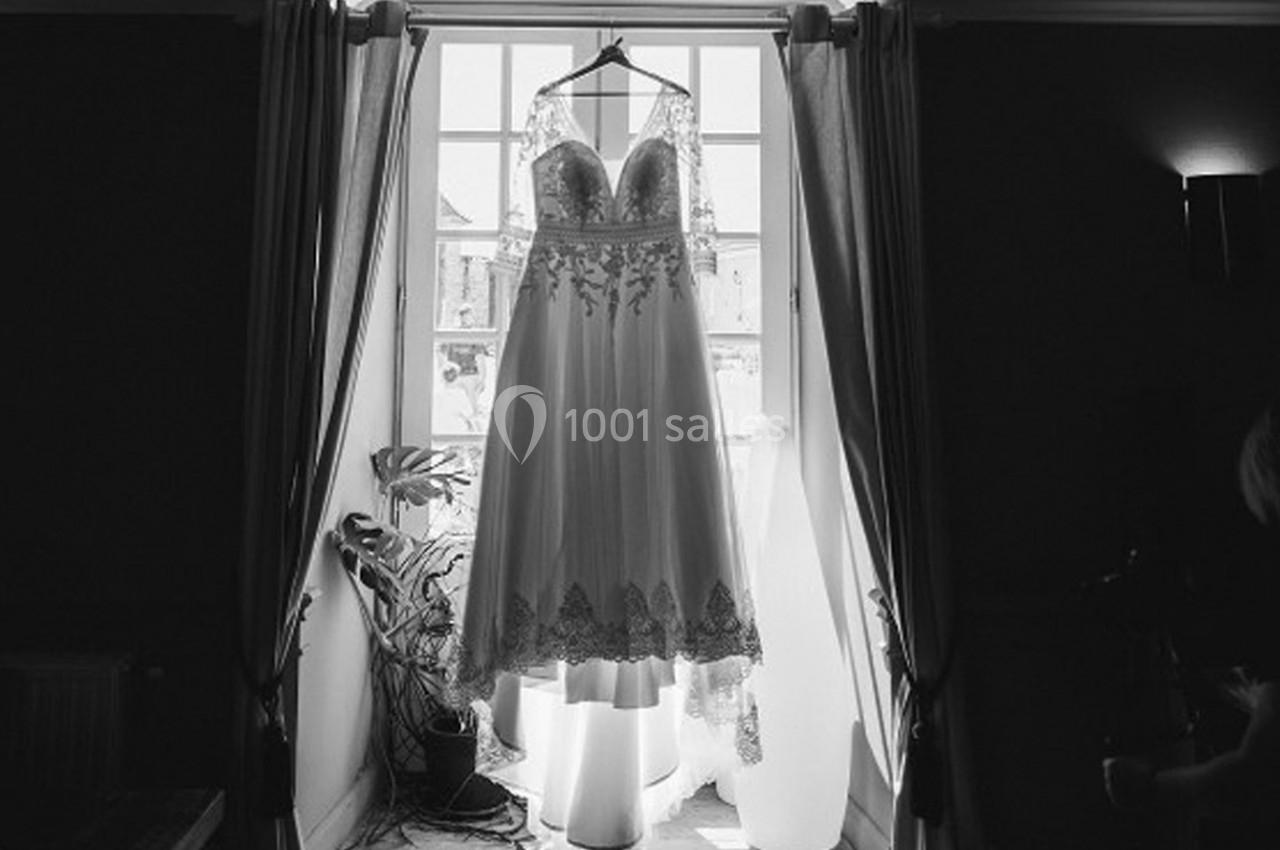 Robe de mariée en dentelle suspendue devant une fenêtre lumineuse avec des rideaux ouverts.