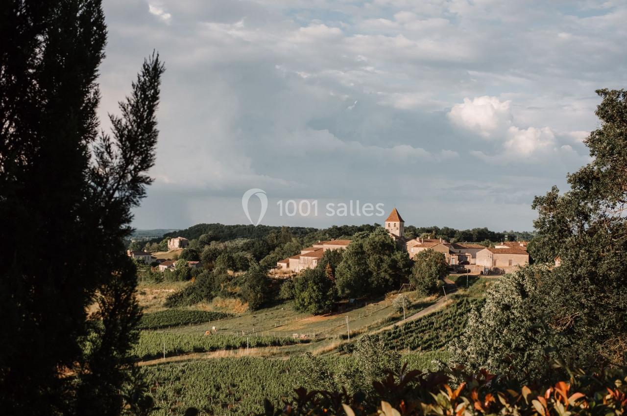 Village rural avec église au clocher pointu, entouré de champs verdoyants et d'arbres sous un ciel partiellement nuageux.