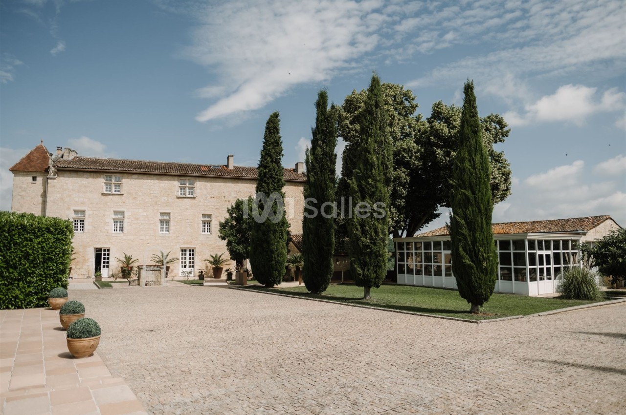 Cour pavée avec un bâtiment en pierre, des cyprès et une véranda entourés de verdure sous un ciel partiellement nuageux.