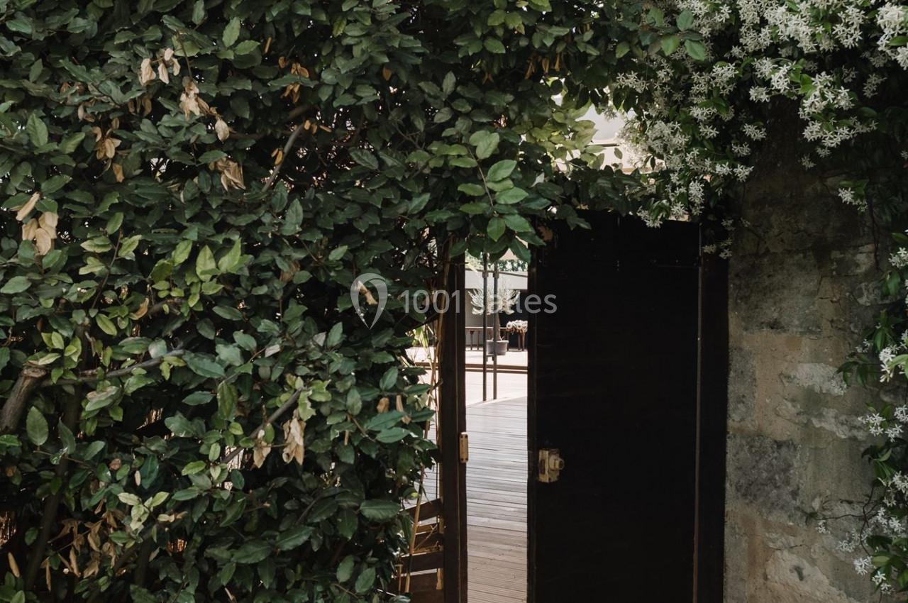 Portail en bois sombre entrouvert, entouré de feuillage dense et de fleurs blanches, donnant sur une terrasse ensoleillée.