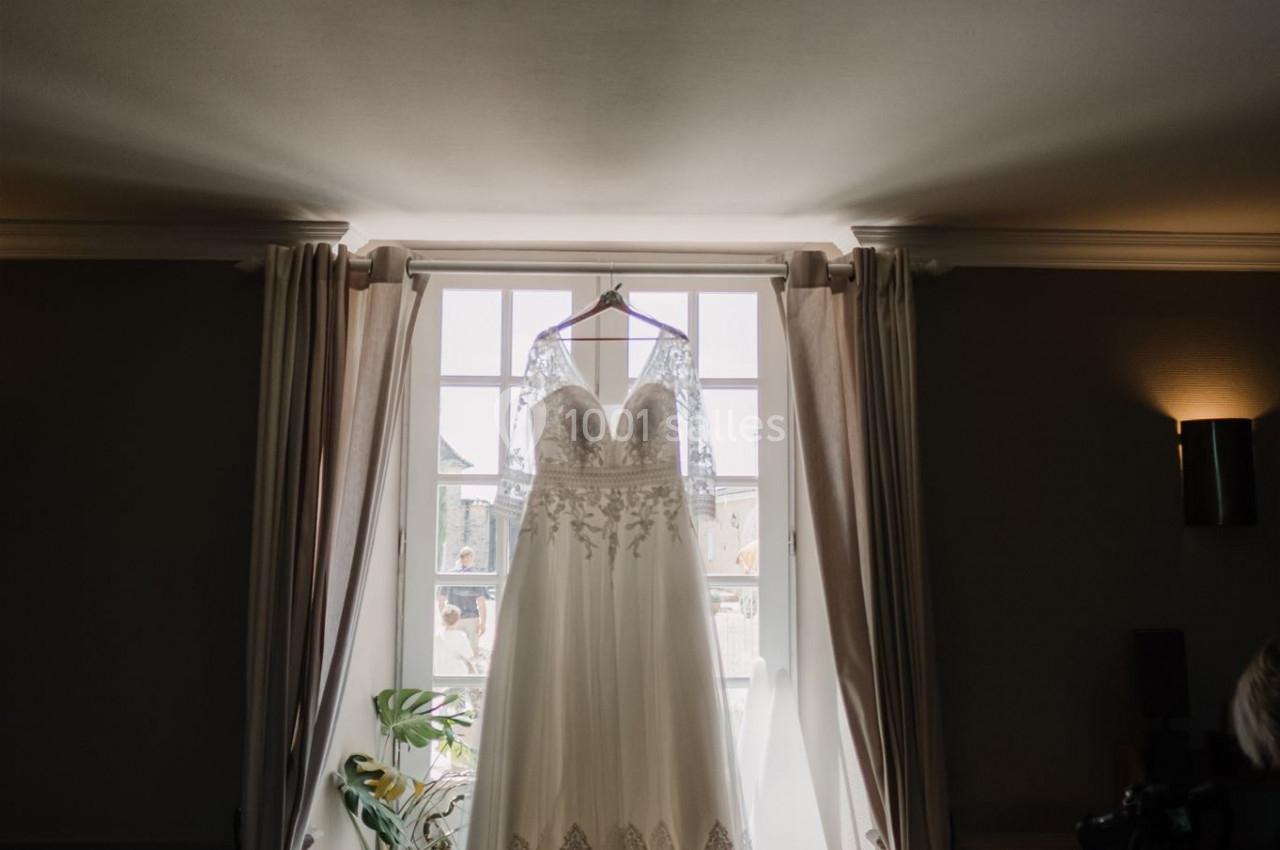 Robe de mariée blanche en dentelle suspendue devant une fenêtre lumineuse avec des rideaux ouverts.