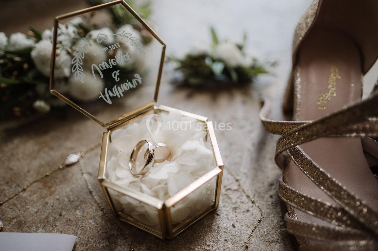 Alliances dorées dans une boîte hexagonale en verre et métal doré, entourée de fleurs et de chaussures scintillantes.