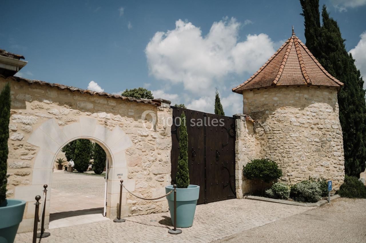 Mur en pierre avec une arche, une tour ronde et un portail en bois, entouré de plantes et de pots bleus.