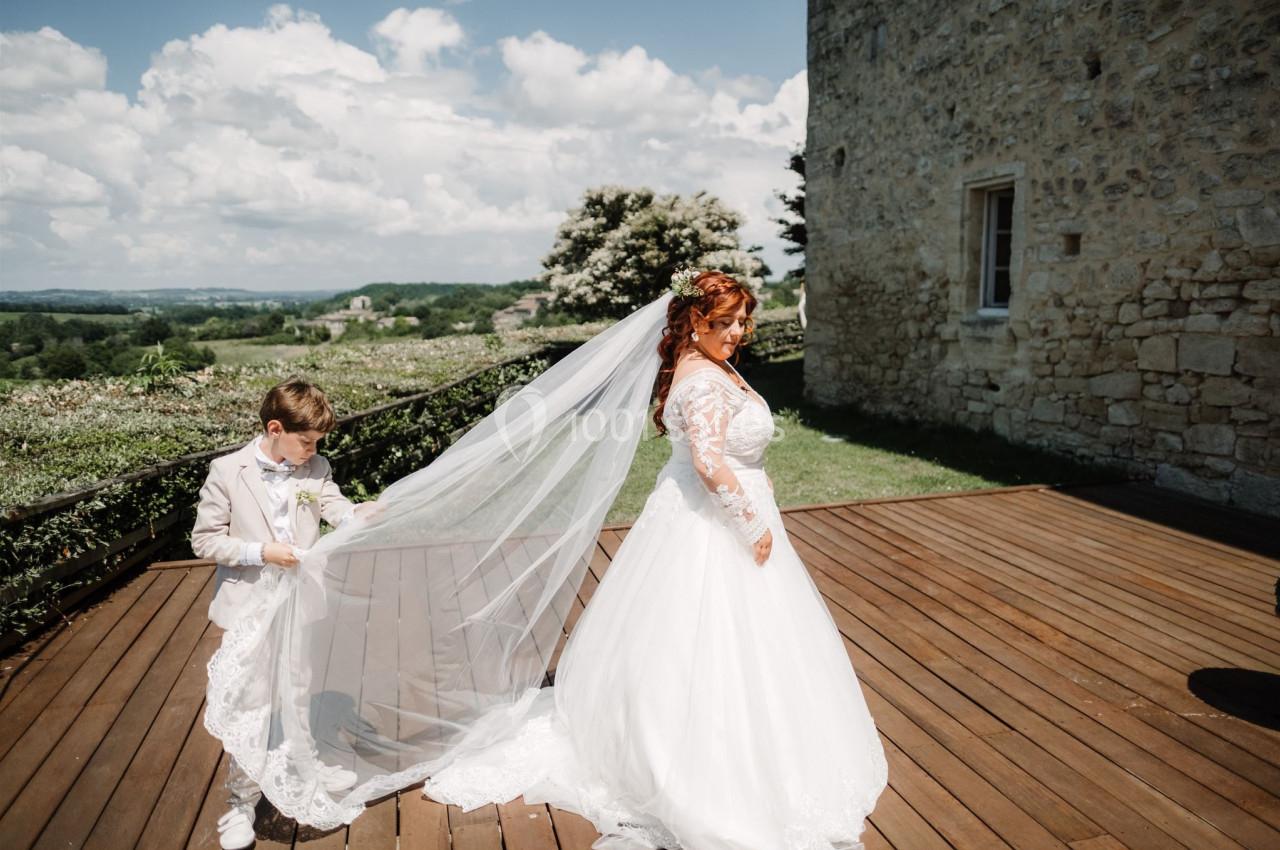 Une mariée en robe blanche marche sur une terrasse en bois, un enfant ajuste son voile sous un ciel dégagé.