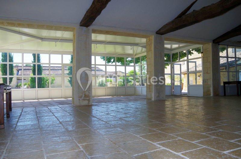 Salle lumineuse avec sol en pierre, poutres apparentes et grandes baies vitrées donnant sur un jardin arboré.