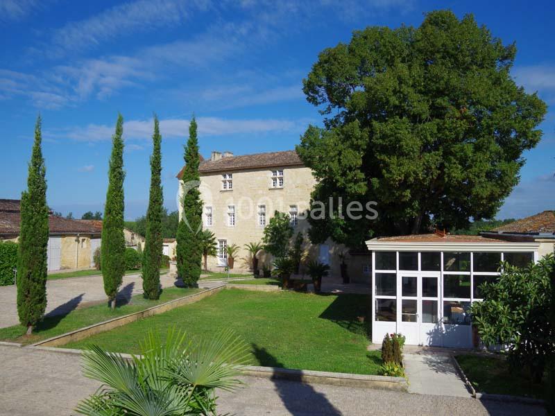 Cour d'un domaine avec un bâtiment en pierre, des cyprès alignés, un grand arbre et une véranda blanche.