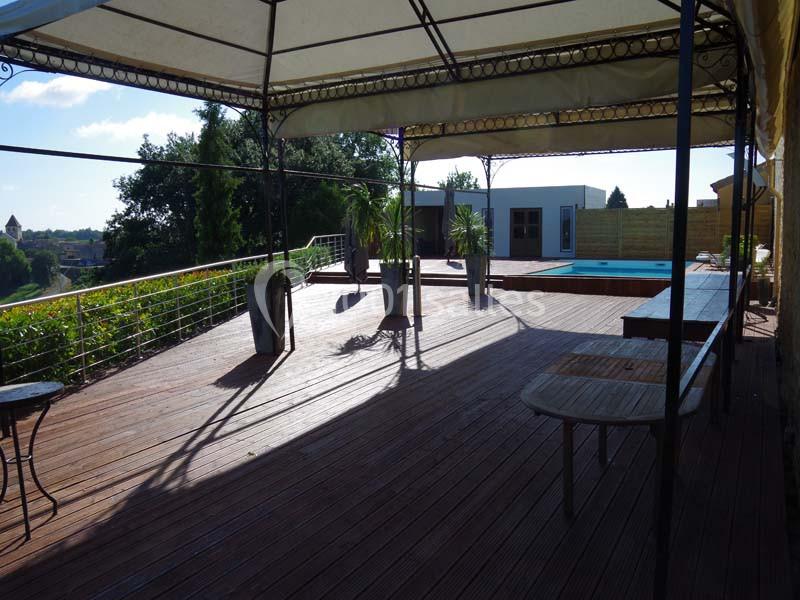 Terrasse en bois avec pergola, vue sur une piscine et un jardin arboré sous un ciel dégagé.