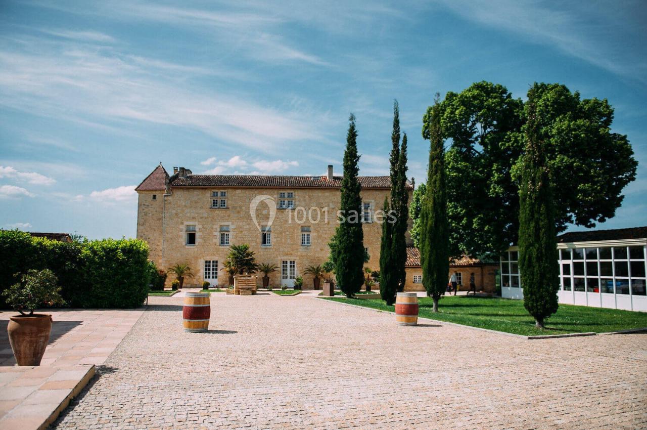 Cour pavée devant un bâtiment en pierre de style ancien, entouré de cyprès et d'arbres sous un ciel dégagé.