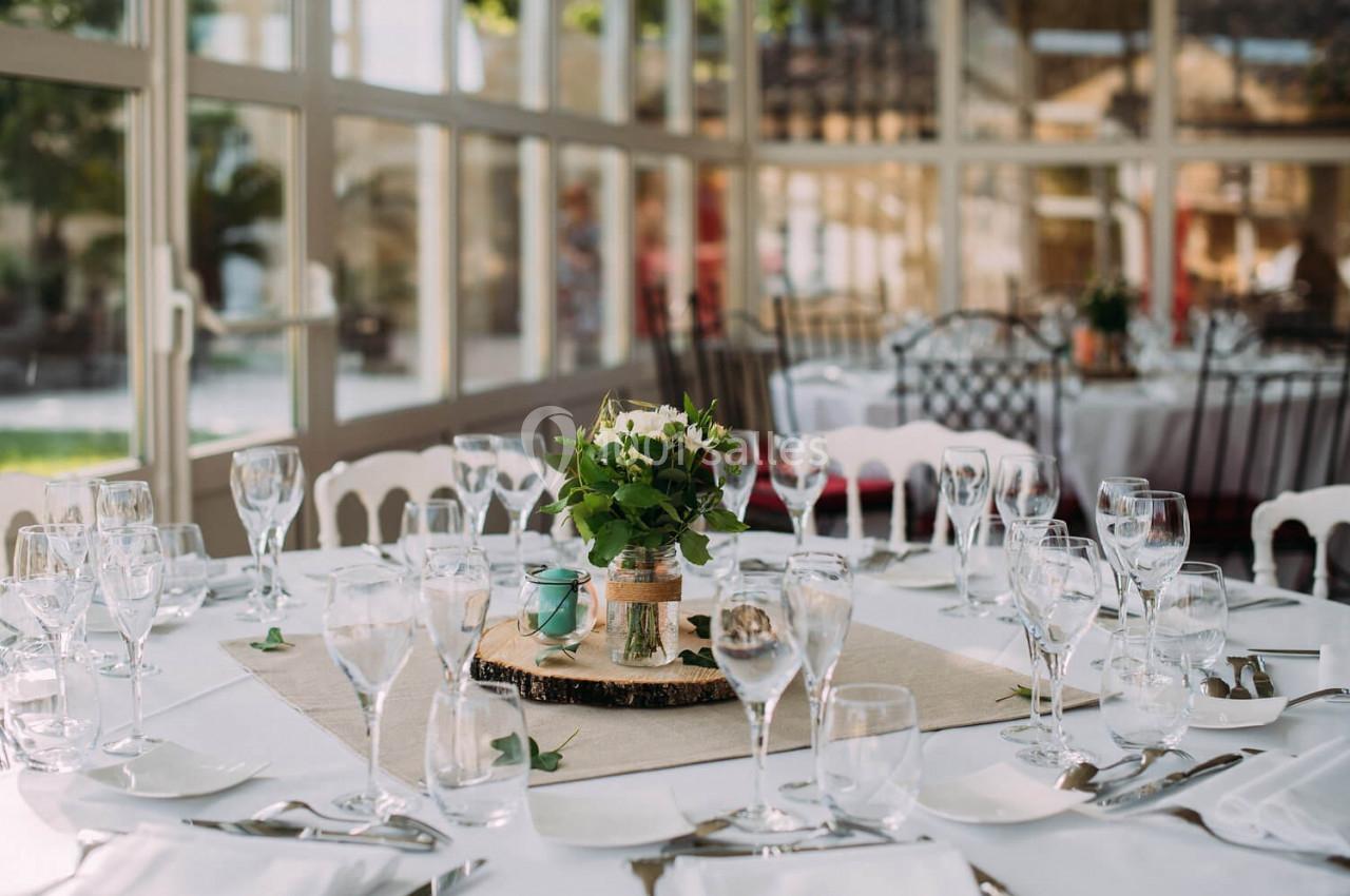 Table dressée avec nappes blanches, verres, couverts et centre de table floral dans une salle lumineuse avec baies vitrées.
