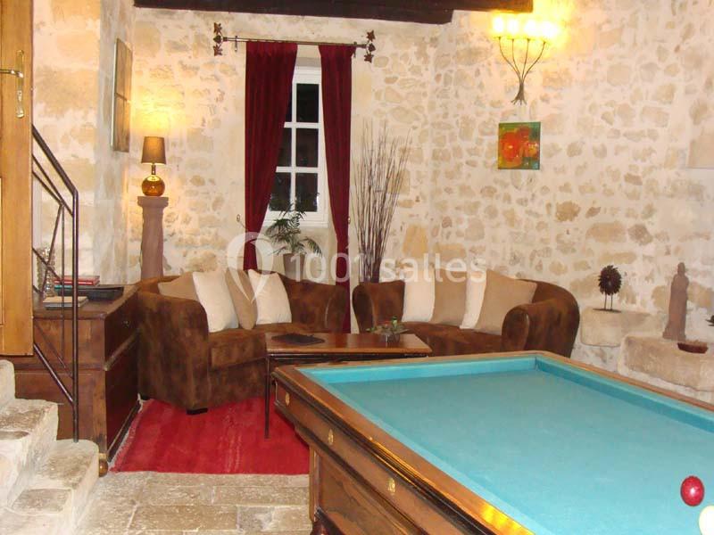 Salon chaleureux avec murs en pierre, canapé en velours, table basse, billard et rideaux rouges devant une fenêtre.