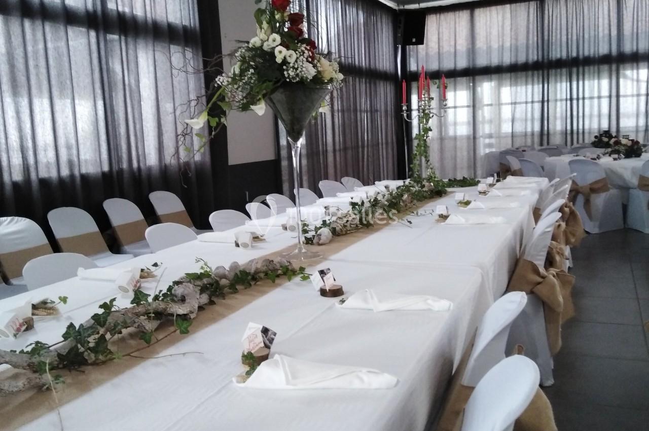 Salle décorée pour un événement avec une longue table dressée, nappes blanches, chaises ornées et centres de table fleuris.