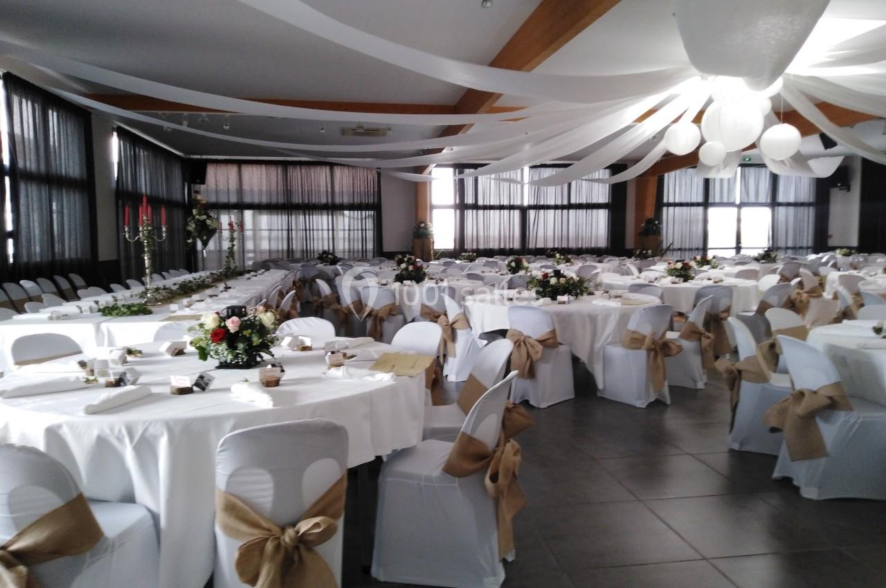 Salle de réception décorée avec des tables rondes, nappes blanches, chaises ornées de nœuds en toile de jute et guirlandes…