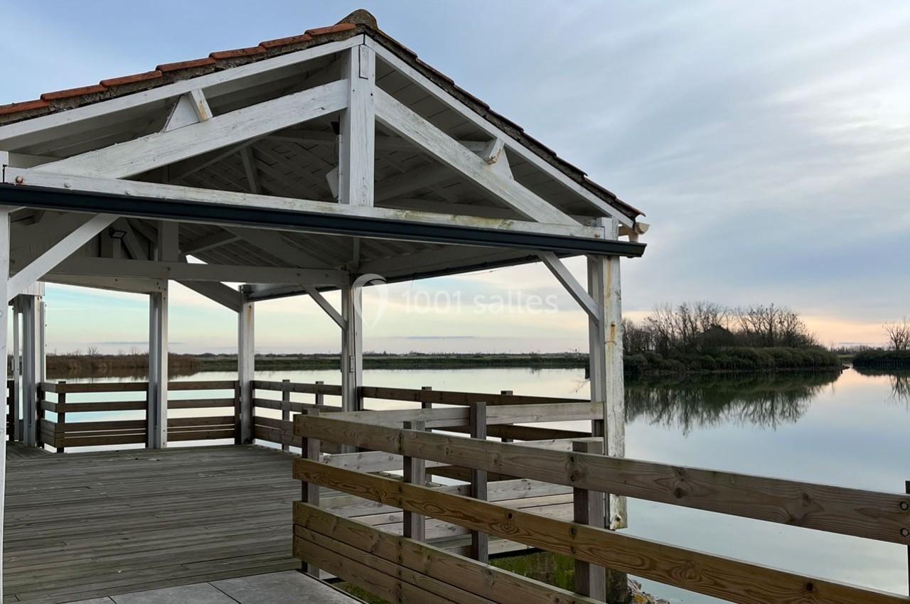 Abri en bois avec vue sur un plan d'eau calme et des arbres en arrière-plan, sous un ciel légèrement nuageux.