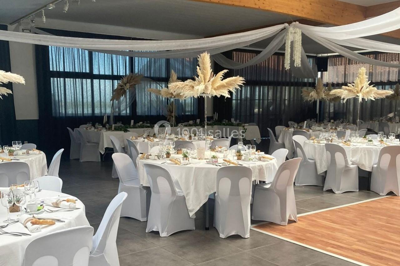 Salle de réception décorée avec des tables rondes, nappes blanches, chaises houssées et ornements de pampas suspendus.