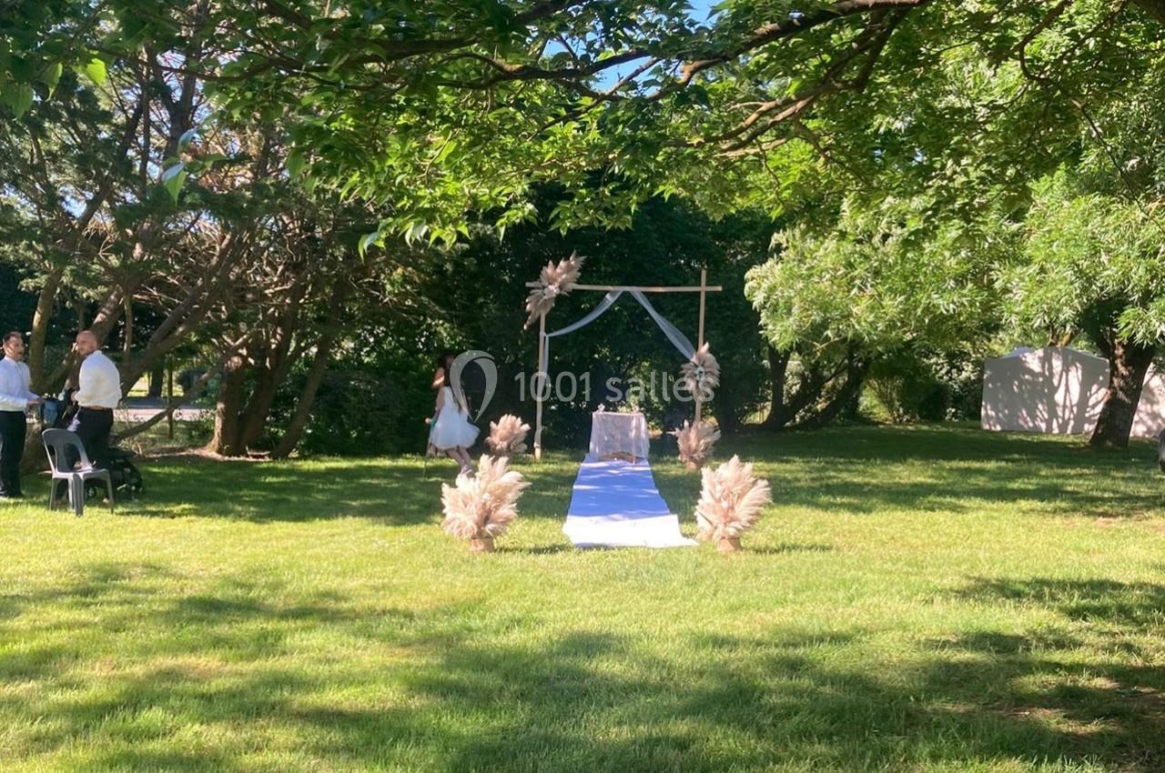 Allée décorée pour une cérémonie en plein air, avec arche florale et chaises sur une pelouse ombragée.