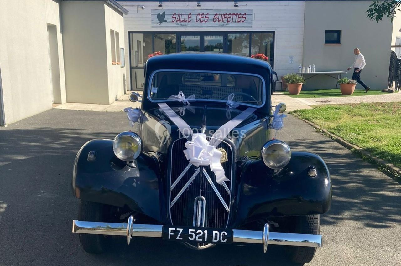 Voiture ancienne noire décorée pour un mariage, stationnée devant une salle des fêtes sous un ciel ensoleillé.