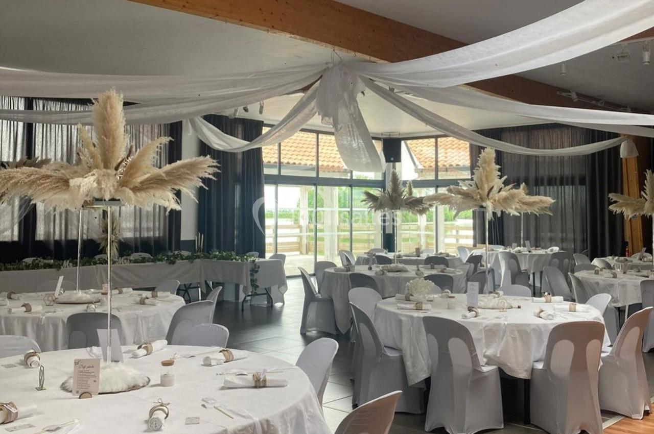 Salle de réception décorée avec des tables rondes, nappes blanches, chaises grises et ornements de plumes.