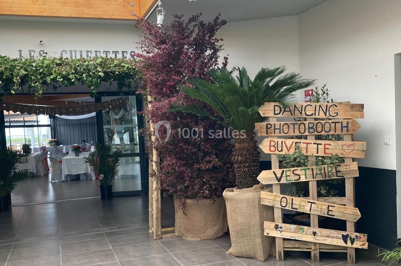 Entrée décorée avec des plantes, un panneau en bois indiquant des activités et une salle aménagée visible en arrière-plan.