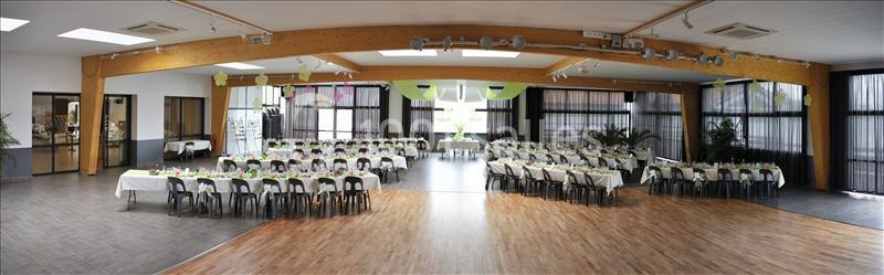 Salle de réception spacieuse avec tables dressées et décorations vertes, éclairée par de grandes baies vitrées.