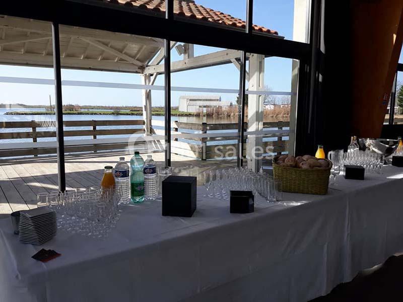 Table dressée avec des verres, boissons et viennoiseries, située près d'une baie vitrée donnant sur un paysage lacustre.