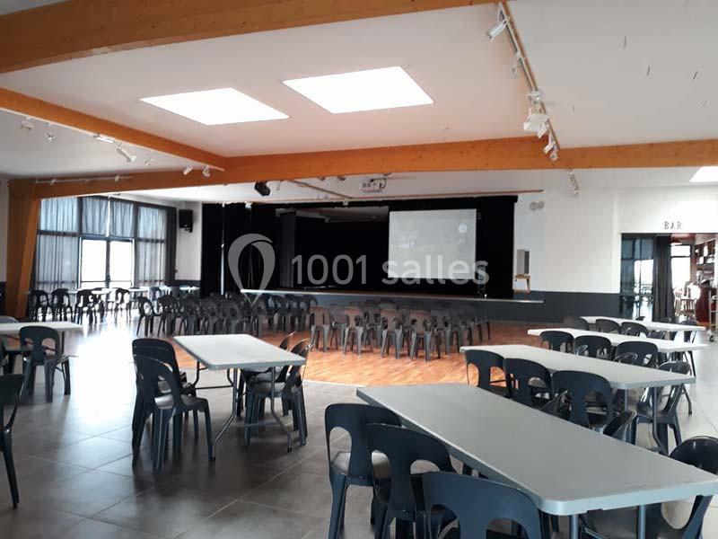 Salle polyvalente avec tables, chaises en plastique et une scène équipée d'un écran de projection.