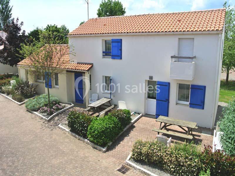 Maison blanche à deux étages avec volets bleus, entourée de végétation et équipée de tables de pique-nique en extérieur.