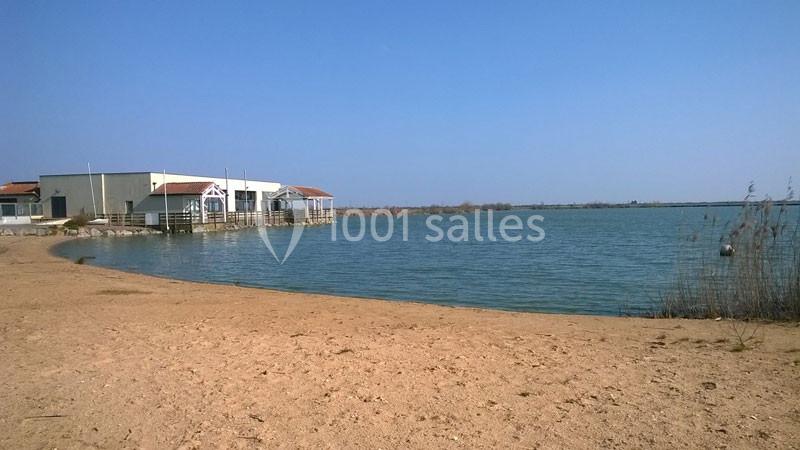 Bâtiments au bord d'un étang calme, avec une plage de sable au premier plan et un ciel dégagé.