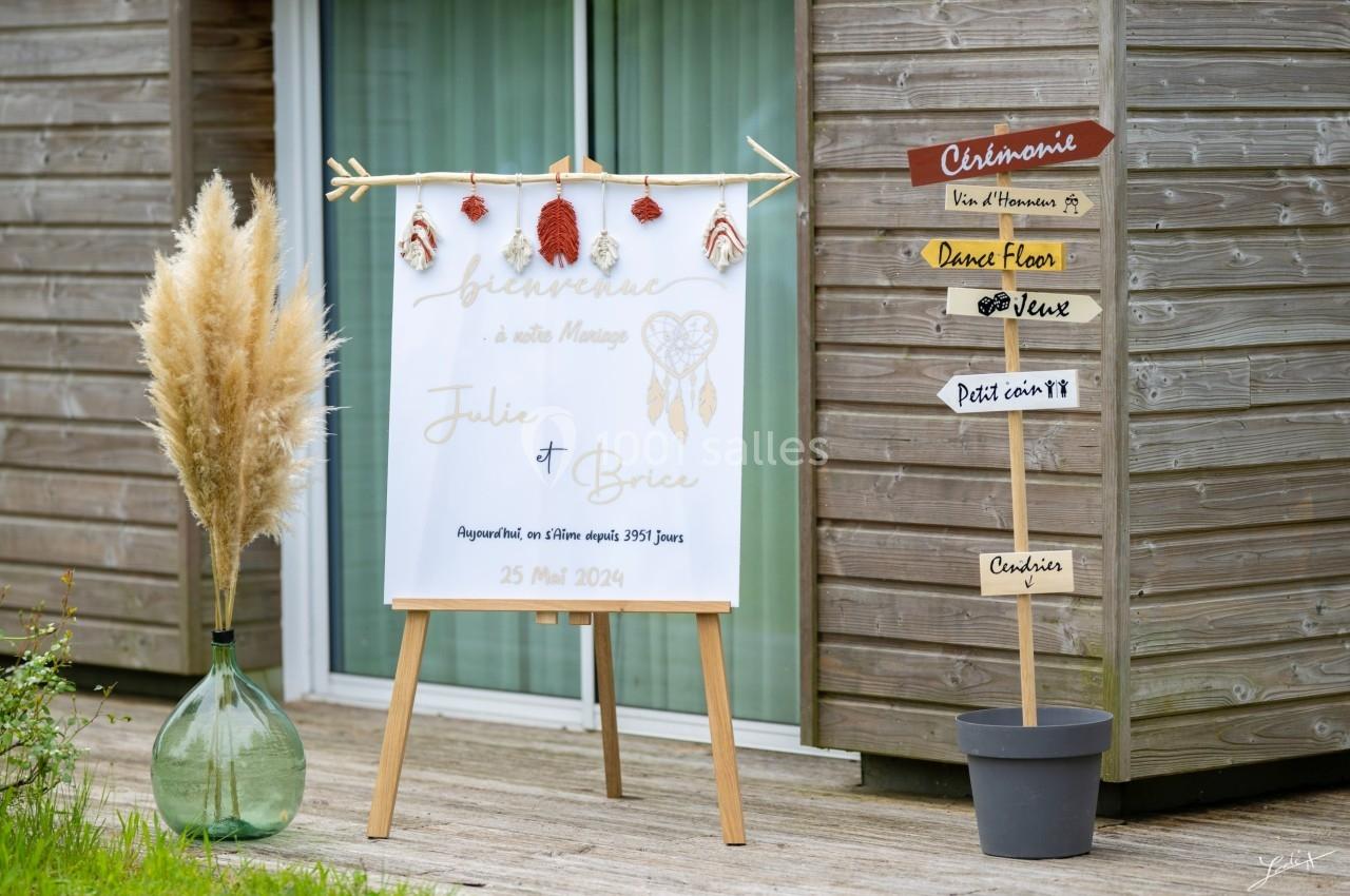 Panneau de bienvenue pour un mariage devant un bâtiment en bois, avec décorations et pancartes directionnelles.