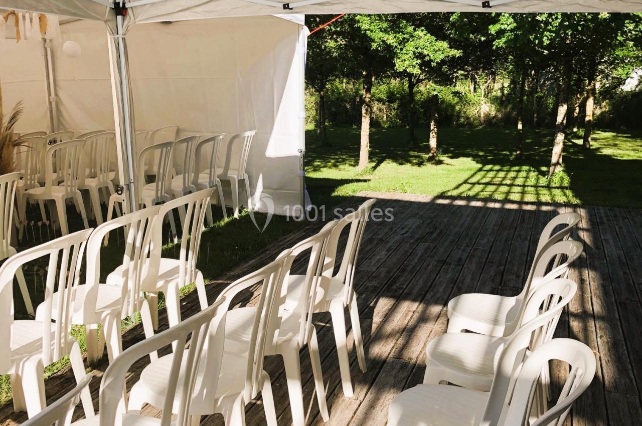 Chaises blanches en plastique alignées sous une tente blanche sur une terrasse en bois, avec un jardin en arrière-plan.