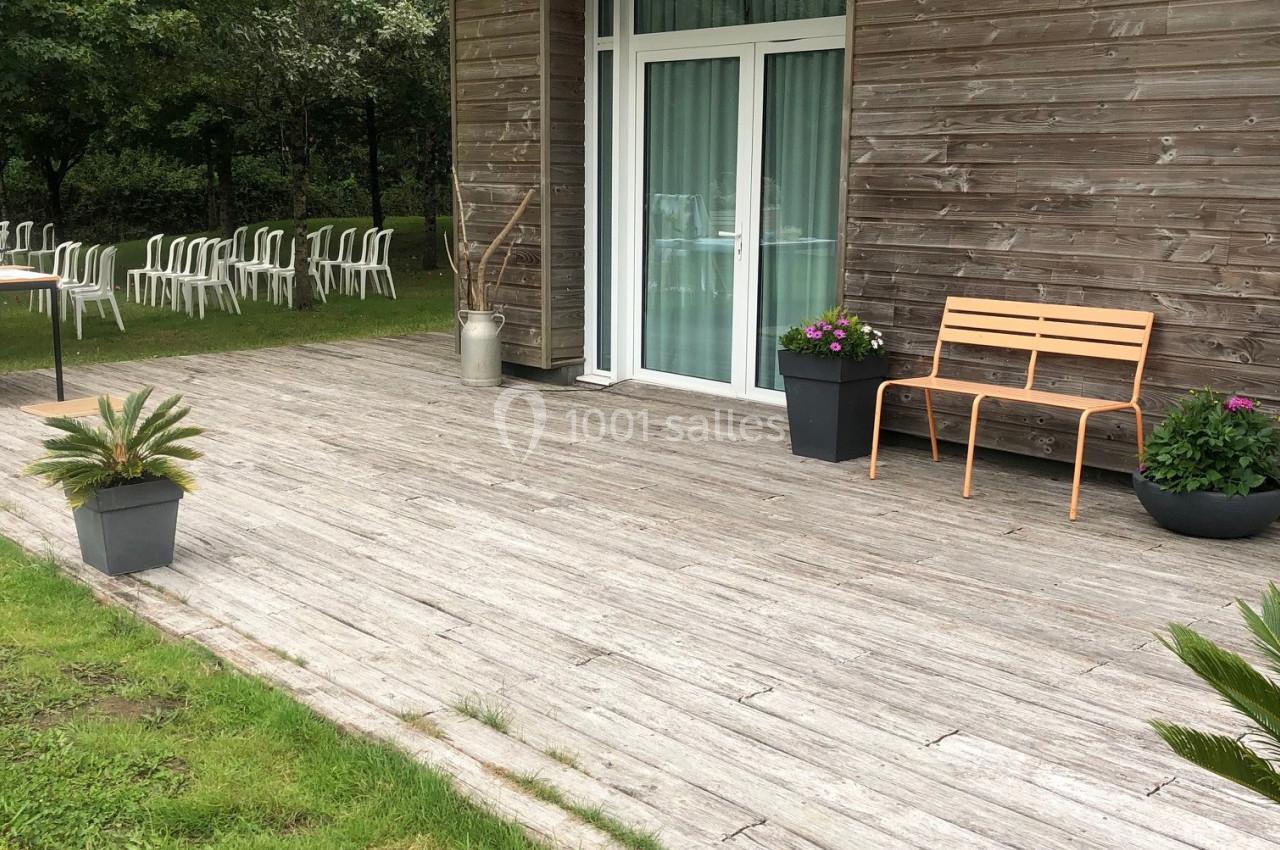 Terrasse en bois avec banc, pots de fleurs et pergola, donnant sur une pelouse et des chaises blanches en arrière-plan.
