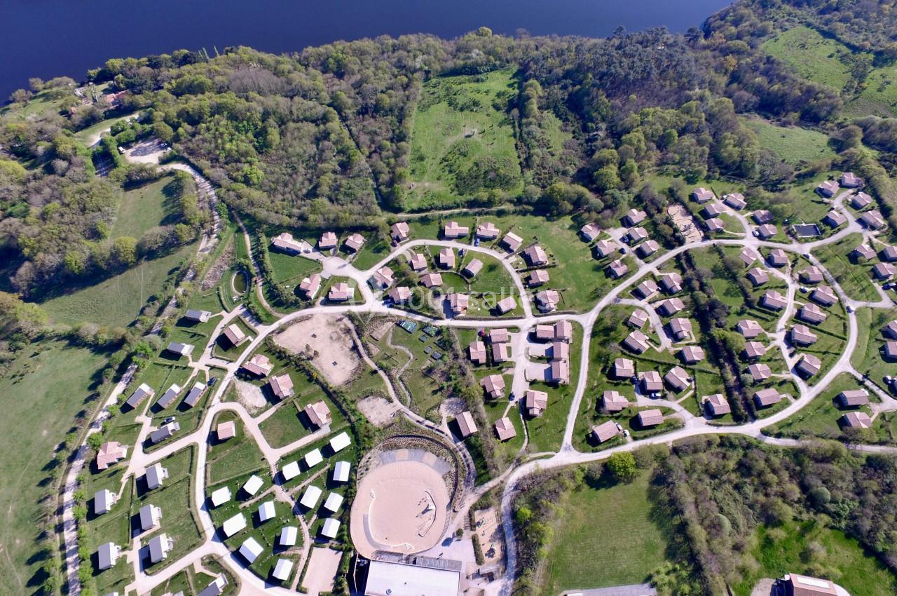 Vue aérienne d'un village avec des maisons dispersées, entouré de verdure et bordé par un cours d'eau.