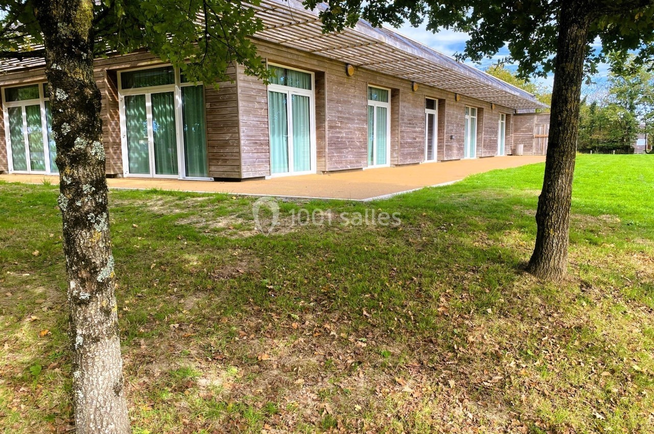 Bâtiment en bois avec de grandes baies vitrées, entouré d'herbe et d'arbres sous un ciel partiellement dégagé.
