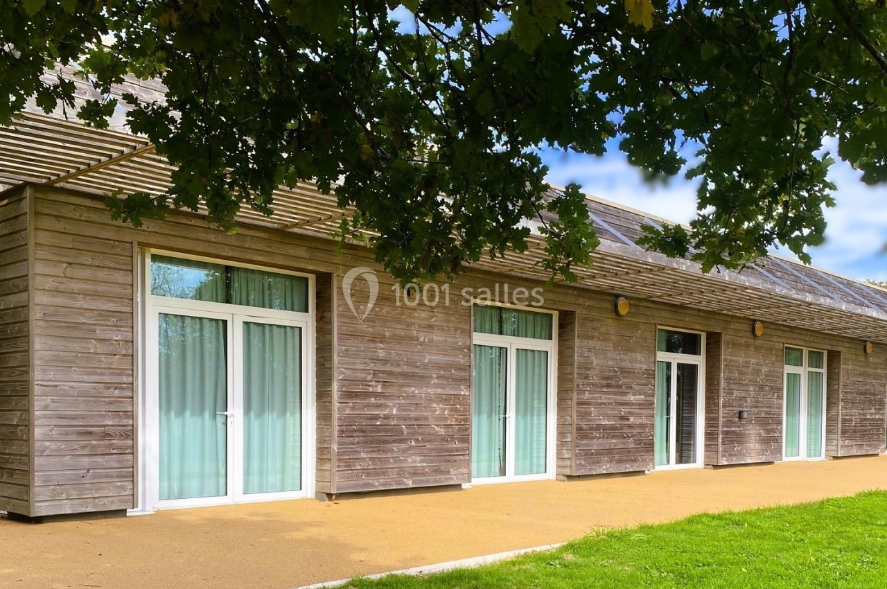 Bâtiment en bois avec de grandes baies vitrées, entouré de verdure et partiellement ombragé par des arbres.