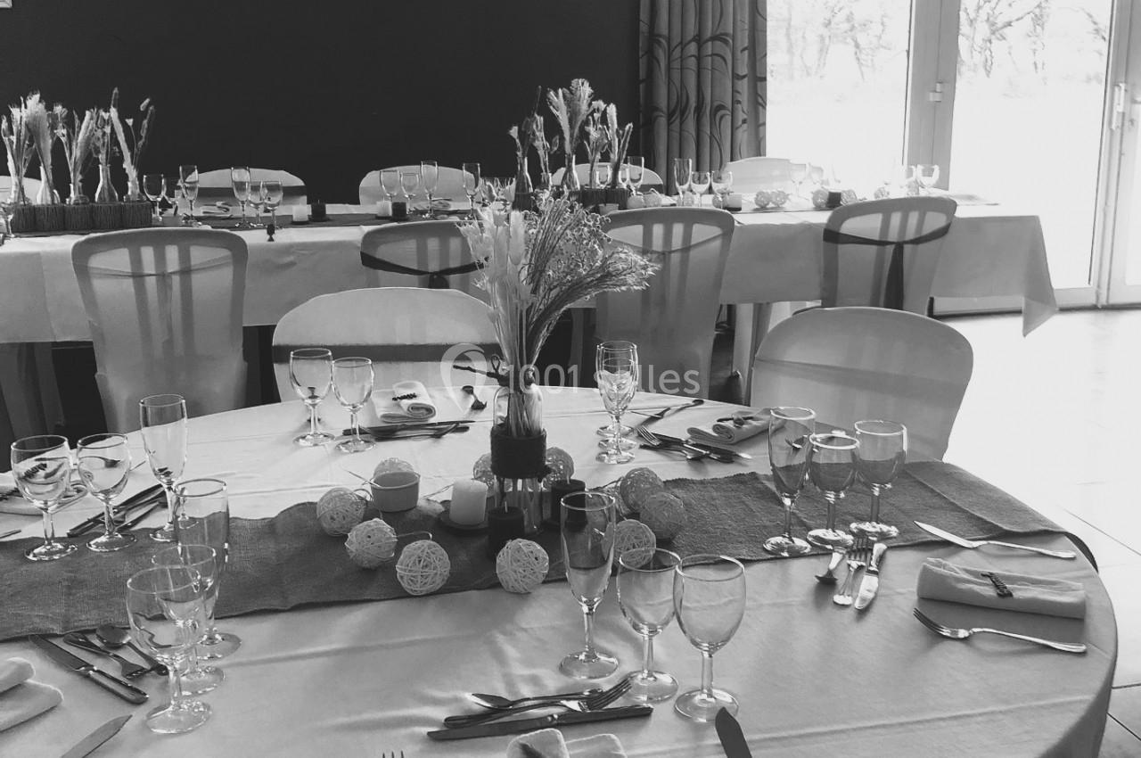 Salle de réception avec tables dressées, nappes blanches, décorations florales et lumière naturelle entrant par une baie…