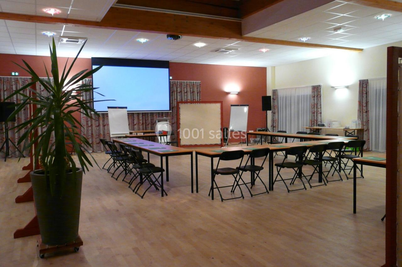 Salle de réunion équipée de tables en U, chaises, projecteur, écrans et plantes décoratives.