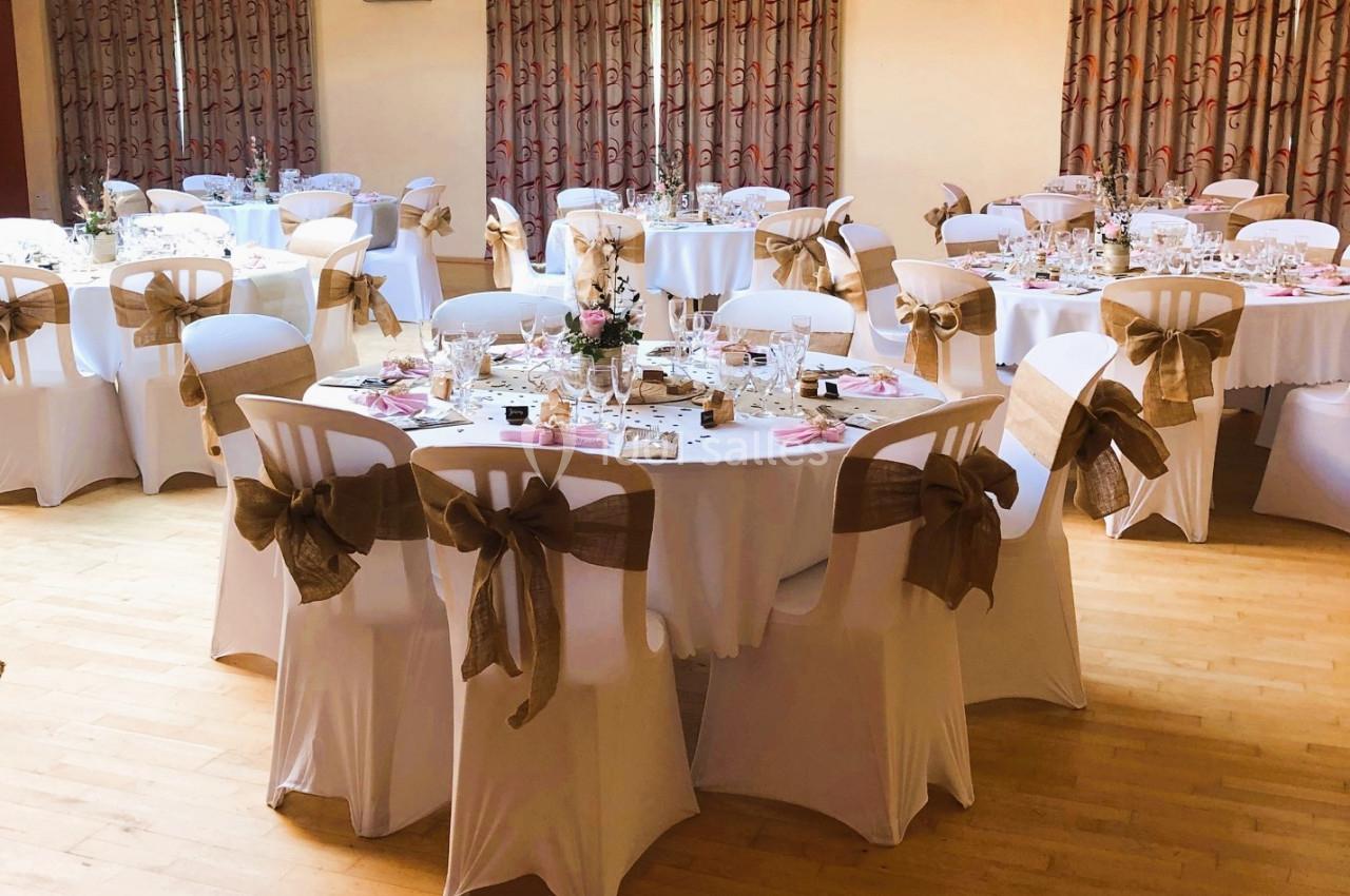 Salle décorée pour un événement avec des tables rondes dressées, nappes blanches et chaises ornées de nœuds beiges.