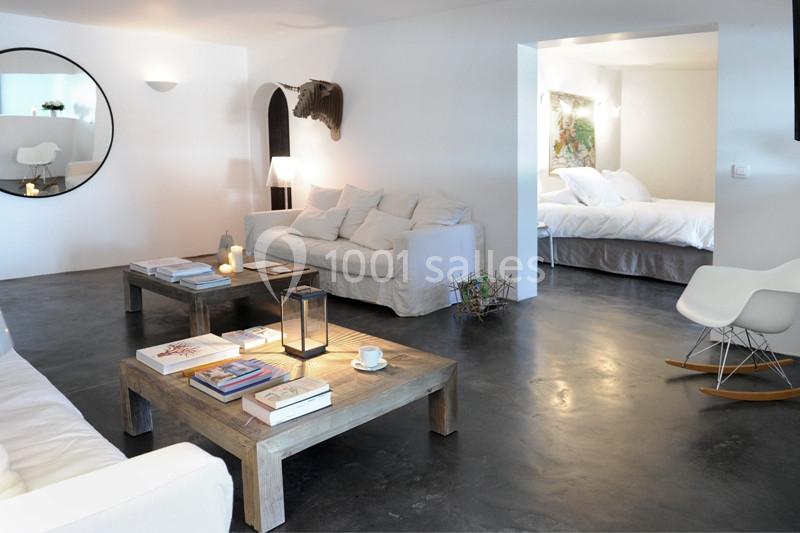 Salon lumineux avec canapé blanc, table basse en bois, livres, bougie allumée, et vue sur une chambre avec lit.