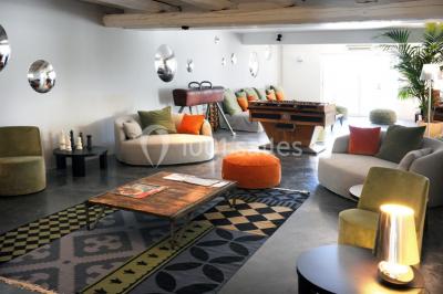 Salon lumineux avec canapé, table basse, tapis à motifs, et chambre en mezzanine avec lit double en arrière-plan.