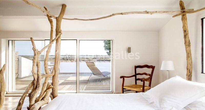 Chambre lumineuse avec lit blanc, chaises en bois, décor rustique et grande baie vitrée donnant sur une terrasse.