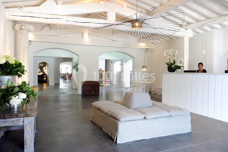 Intérieur lumineux avec poutres apparentes, canapé beige au centre, réceptionniste à un comptoir blanc en arrière-plan.