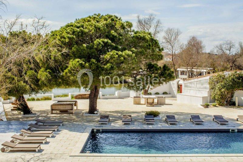 Piscine extérieure entourée de transats, arbres et végétation, avec vue sur un plan d'eau et une terrasse pavée.