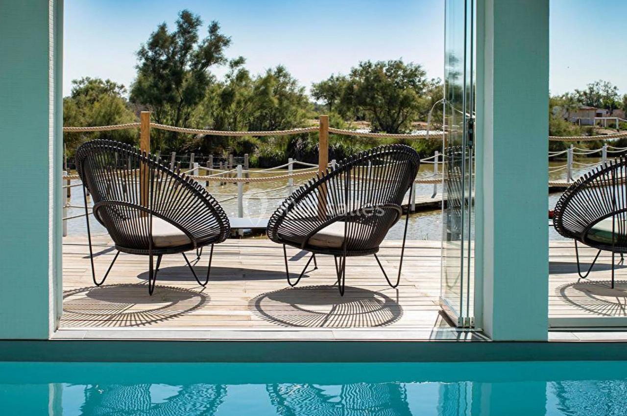 Deux fauteuils en rotin sur une terrasse en bois surplombant un plan d'eau, vus depuis une piscine intérieure.
