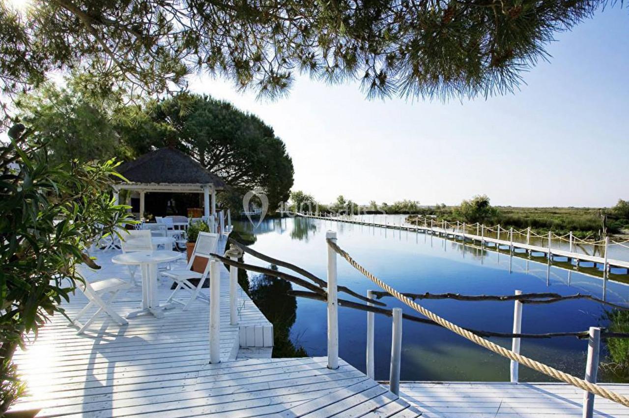 Terrasse en bois blanche bordant un étang calme, avec des tables, des chaises et un kiosque sous des arbres.