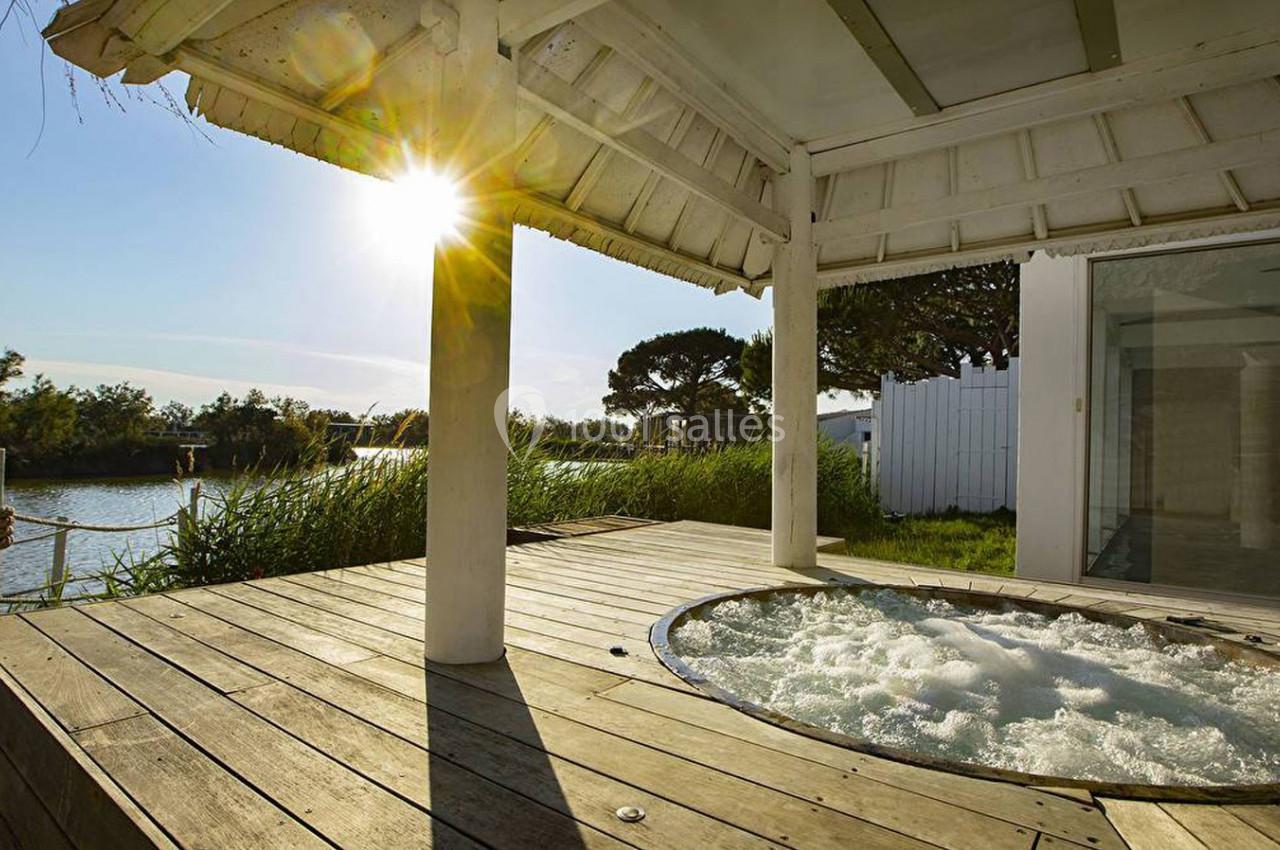 Terrasse en bois avec un jacuzzi encastré, abritée par une pergola, près d'un plan d'eau sous un soleil éclatant.
