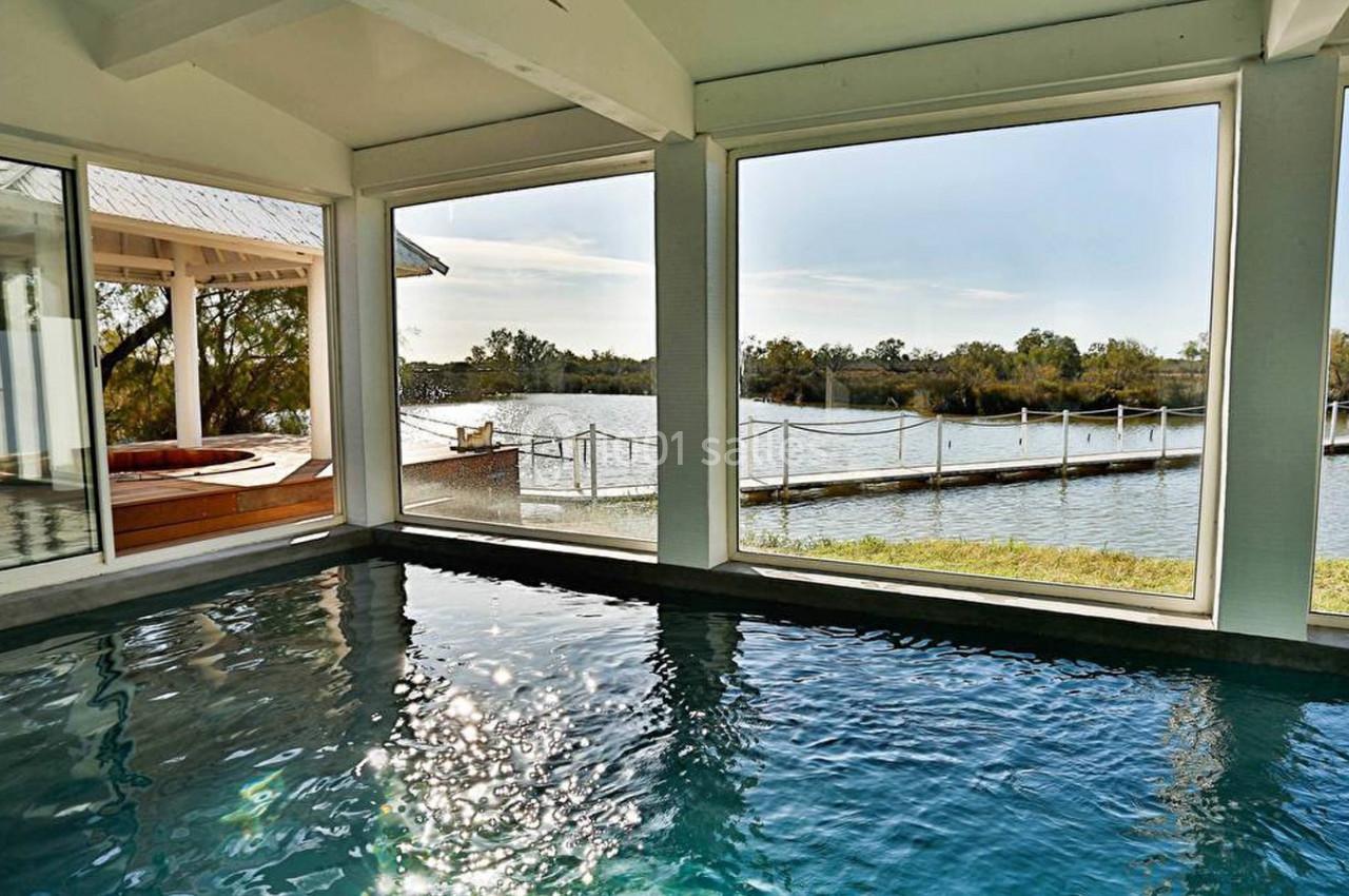 Vue intérieure d'une piscine donnant sur un paysage de lac avec ponton et végétation à travers de grandes baies vitrées.