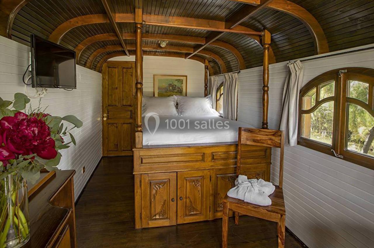 Intérieur d'une roulotte en bois aménagée avec un lit, une chaise, des fenêtres cintrées et un bouquet de fleurs.