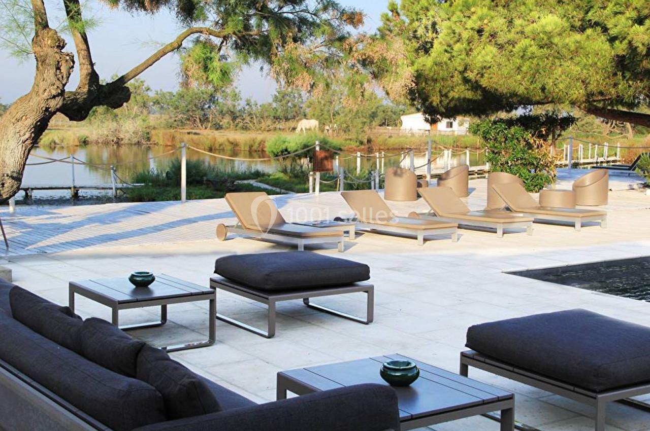 Terrasse avec mobilier de jardin moderne, transats et tables basses, près d'un plan d'eau entouré de végétation.