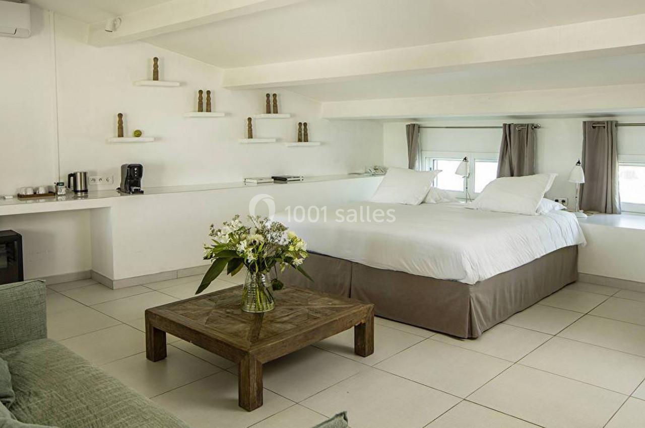 Chambre lumineuse avec lit double, table basse en bois, canapé vert et étagères décoratives sur un mur blanc.